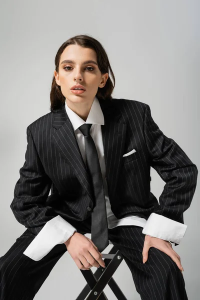 Brunette woman in white shirt and black striped suit looking at camera while sitting isolated on grey — Stockfoto