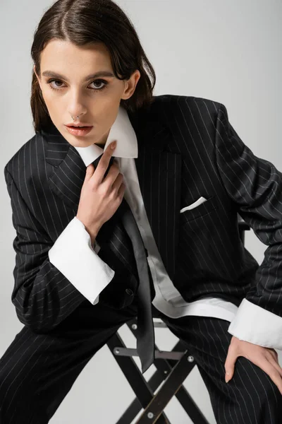 Молода жінка в білій сорочці і чорний смугастий блейзер торкається краватки, сидячи і дивлячись на камеру ізольовано на сірому — стокове фото