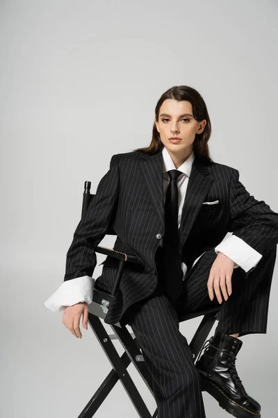 Стильна жінка в модному наряді, дивлячись на камеру, позує на стілець ізольовано на сірому — стокове фото