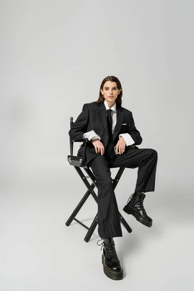 Полная длина модной женщины в грубых сапогах и черном огромном костюме, сидящей на стуле и смотрящей на камеру на сером фоне — стоковое фото