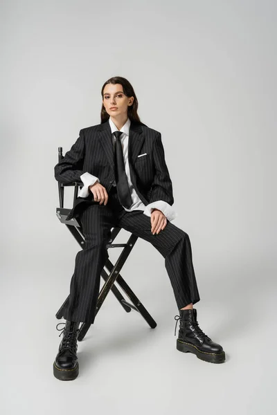 Full length of trendy woman in oversize formal wear and laced-up boots looking at camera while sitting on grey — Foto stock