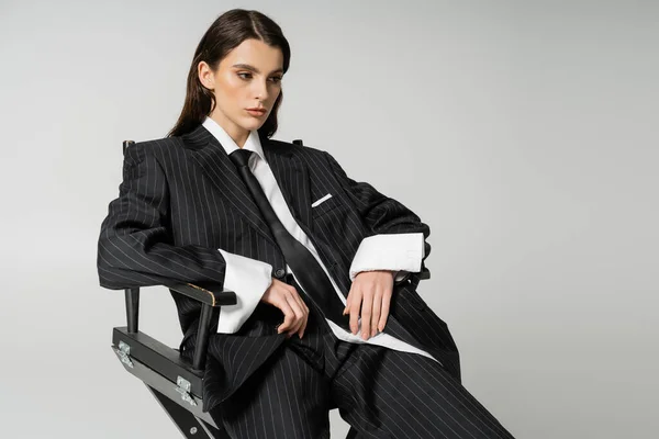 Pretty brunette woman in white shirt and black striped suit sitting isolated on grey - foto de stock