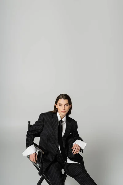 Модна жінка в обмеженому формальному одязі сидить і дивиться на камеру ізольовано на сірому з копіювальним простором — стокове фото
