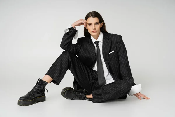 Full length of young model in black elegant pantsuit and rough boots sitting with hand near head on grey background — Stockfoto