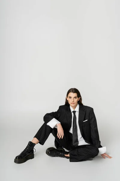 Full length of fashionable woman in black elegant suit and rough boots sitting on grey background - foto de stock