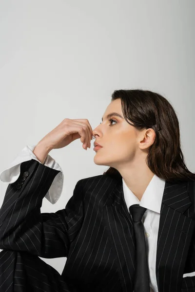 Trendy woman in white shirt and black blazer with tie holding hand near face and looking away isolated on grey - foto de stock