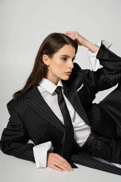 Stylish woman in black striped suit and white oversize shirt posing with hand near head on grey background — Foto stock
