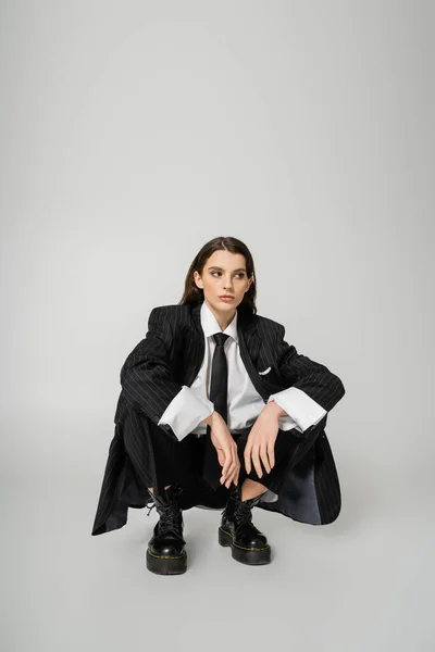 Young model in black oversize suit and rough boots sitting on haunches and looking away on grey background - foto de stock