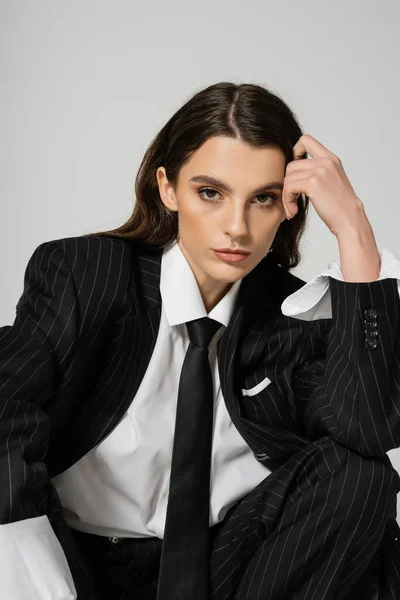 Pretty woman in white shirt and black blazer with tie holding hand near head and looking at camera isolated on grey - foto de stock