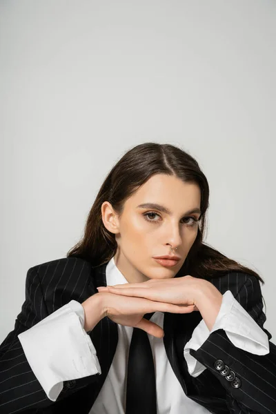 Portrait of brunette woman in stylish attire posing with hands under chin while looking at camera isolated on grey — Stockfoto