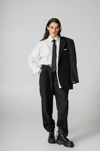 Full length of young model in oversize formal wear and rough boots standing with hand in pocket on grey background — Stockfoto