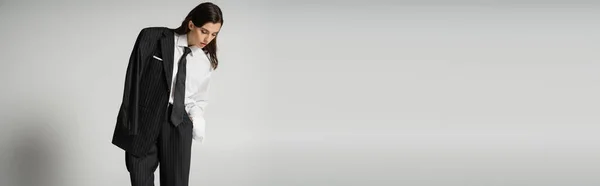 Stylish brunette woman in oversize formal wear standing and looking down on grey background, banner — Fotografia de Stock