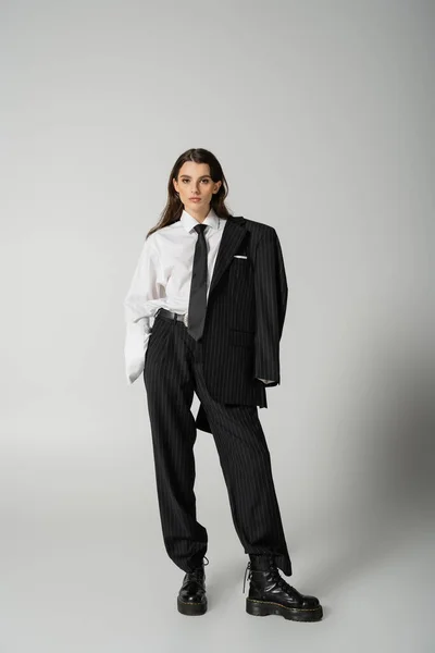 Модна жінка в надмірному формальному одязі і шнурових чоботях, що стоять з рукою в кишені на сірому фоні — стокове фото