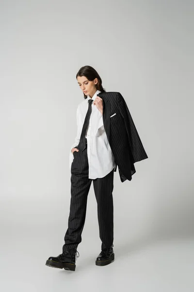 Young model in oversize shirt and black pants holding black blazer while walking in rough boots on grey background - foto de stock