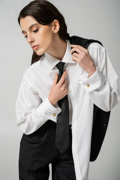 Модна жінка в розмірній сорочці торкається чорної краватки, позує ізольовано на сірому — стокове фото
