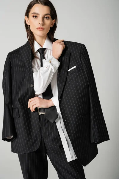 Stylish brunette woman in elegant oversize attire looking at camera isolated on grey — Fotografia de Stock