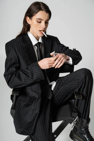 Trendy woman in black oversize suit sitting with cigarette and lighter isolated on grey — Stockfoto