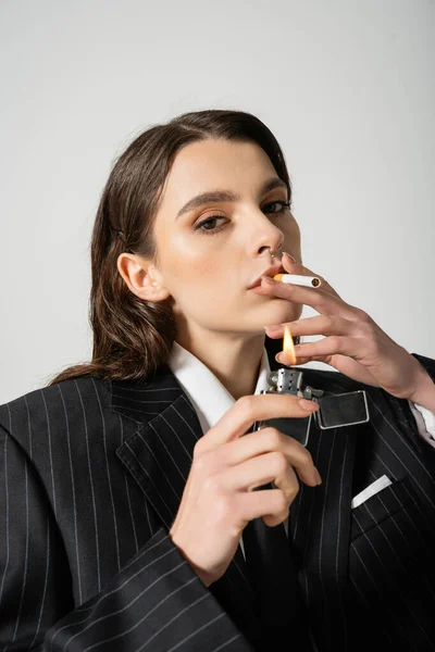 Fashionable woman with makeup and piercing lighting cigarette and looking at camera isolated on grey — Stock Photo