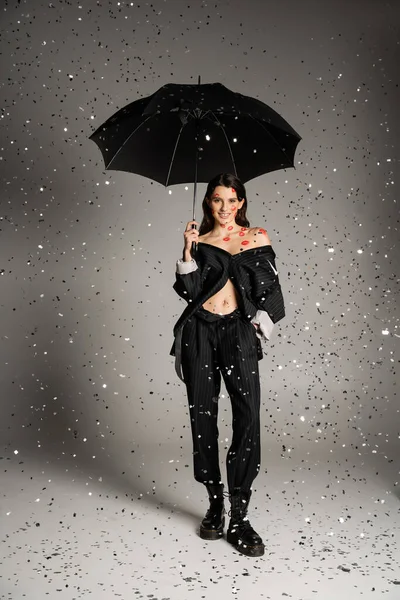 Full length of smiling and stylish woman with red kisses on body standing with black umbrella under falling confetti on grey background — Foto stock