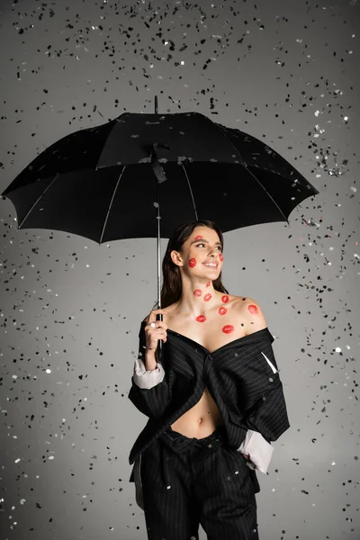 Sexy and stylish woman standing with hand in pocket under black umbrella and shiny confetti on grey background — Stock Photo