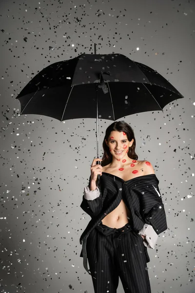 Cheerful woman with red lip prints on body wearing black clothes and standing with umbrella under falling confetti on grey background — Stock Photo