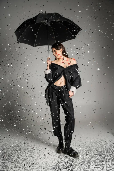 Happy woman in black stylish clothes standing with umbrella under sparkling confetti on grey — Stock Photo