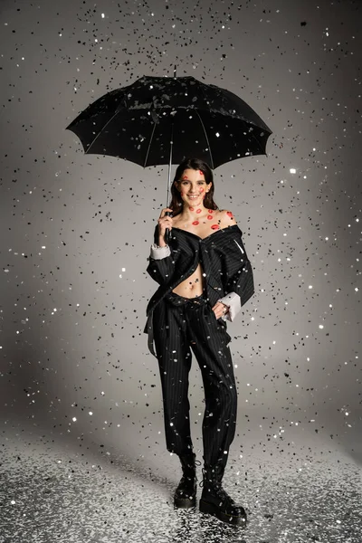 Happy young woman in stylish clothes standing with black umbrella under shiny confetti on grey background — Fotografia de Stock
