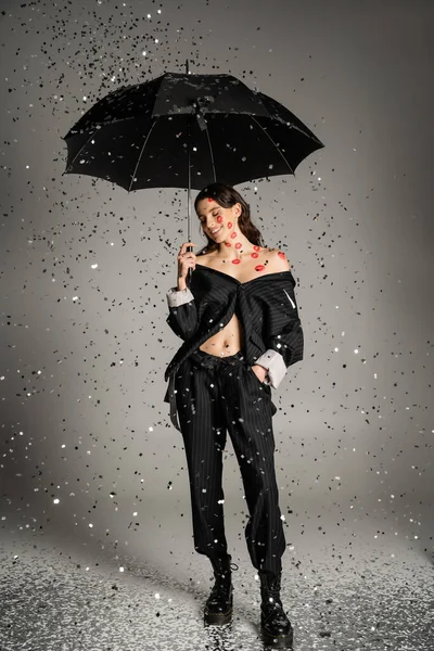 Happy and stylish woman with red lip prints on body standing with black umbrella under shiny confetti on grey background — Foto stock
