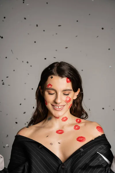 Brunette woman with red kiss prints on face and bare shoulders smiling with closed eyes on grey background - foto de stock