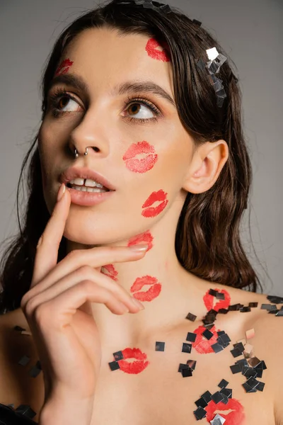 Сексуальна і мрійлива жінка з червоними поцілунками на обличчі і тілі торкається губ і дивиться ізольовано на сірий — стокове фото