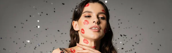 Young brunette woman with kiss prints on face and body posing under falling confetti on grey background, banner — Foto stock