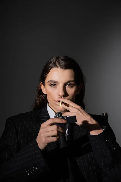 Stylish brunette woman in black blazer lighting cigarette and looking at camera on dark grey background — Stockfoto