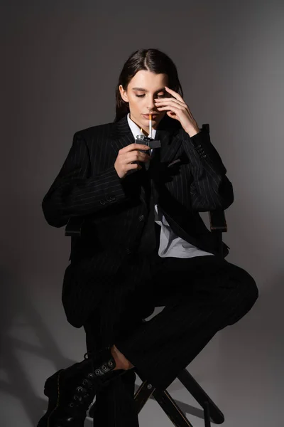 Modische Frau im schwarzen Strumpfanzug zündet sich Zigarette an, während sie auf dunkelgrau sitzt — Stockfoto