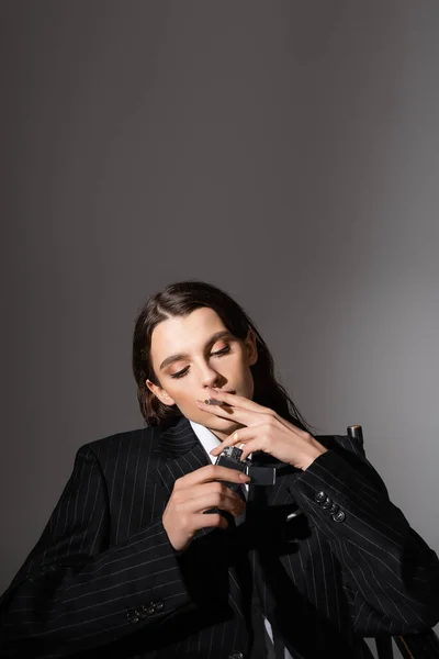 Стильна брюнетка в чорному костюмі сидить і освітлює сигарету на сірому з копіювальним простором — стокове фото