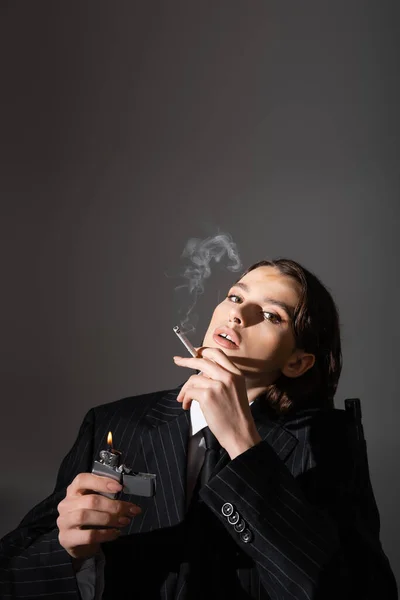 Sensual woman in black striped suit holding lighter and smoking isolated on grey with copy space — Stock Photo