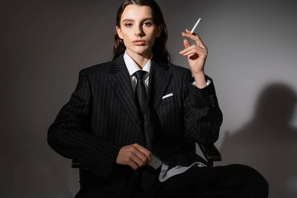 Brunette woman in elegant suit sitting with cigarette and looking at camera on dark background with shadow — Photo de stock