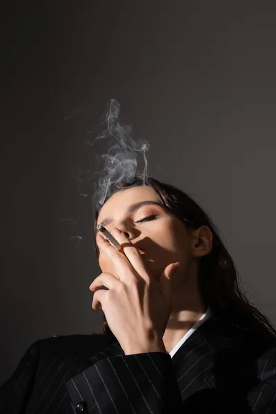 Young woman in black and striped blazer smoking with closed eyes isolated on grey with copy space — Photo de stock