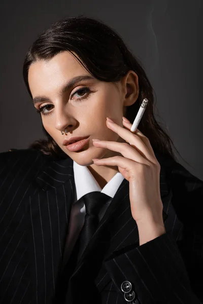 Портрет брюнетки в черном полосатом блейзере, держащей сигарету и смотрящей на камеру, изолированную на темно-сером — стоковое фото