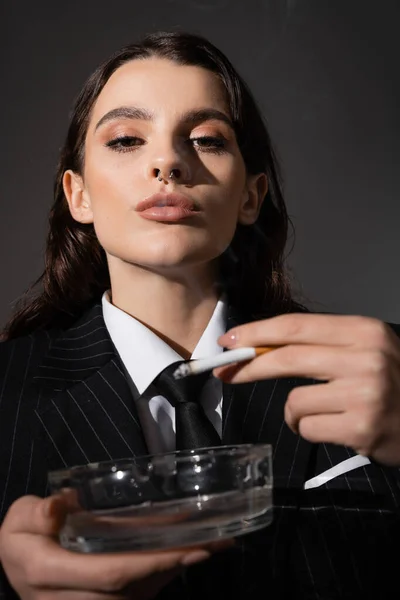 Portrait of fashionable woman with makeup and piercing holding ashtray and cigarette isolated on dark grey - foto de stock