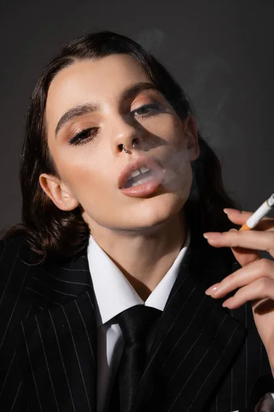 Портрет элегантной женщины с макияжем и пирсингом, смотрящей в камеру во время курения, изолированного на темно-сером — стоковое фото