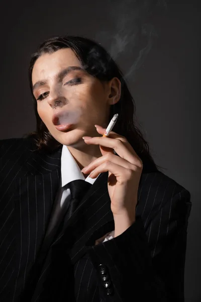 Sensual brunette woman in black blazer and tie smoking isolated on dark grey — Foto stock