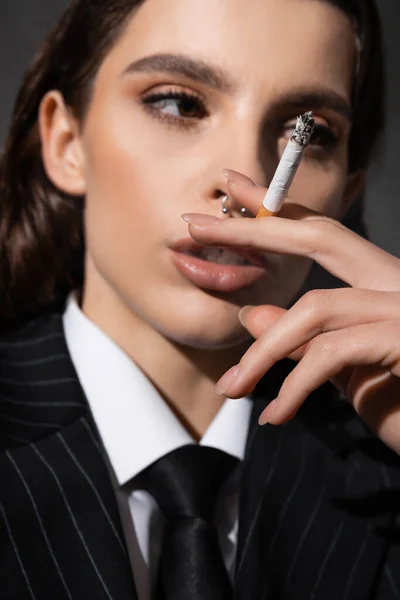 Portrait of stylish blurred woman with makeup and piercing holding cigarette isolated on dark grey - foto de stock