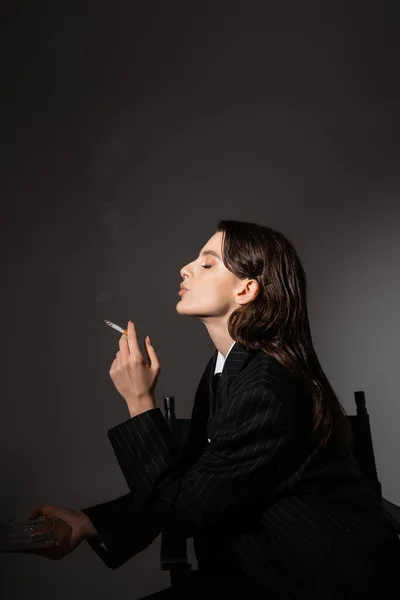 Вид сбоку брюнетки в черном блейзере, держащей пепельницу и курящей на темно-сером фоне — стоковое фото