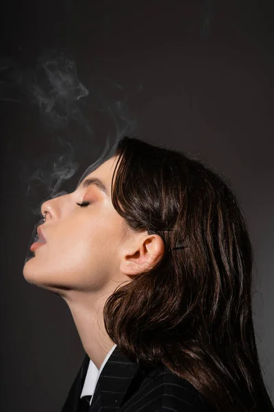 Profile of stylish brunette woman with makeup and piercing smoking isolated on dark grey — Stockfoto