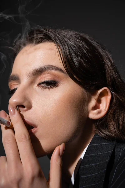 Portrait of stylish brunette woman smoking and looking at camera on black background — Photo de stock