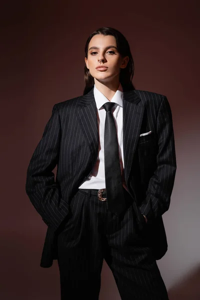Trendy woman in black striped pantsuit and tie standing with hands in pockets and looking at camera on brown background — Foto stock