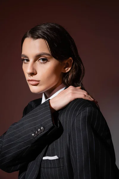 Portrait of elegant brunette woman in black and striped blazer posing with hand on shoulder on brown — Foto stock