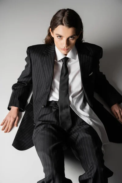 High angle view of fashionable model in oversize shirt and black pantsuit sitting on grey background — Foto stock