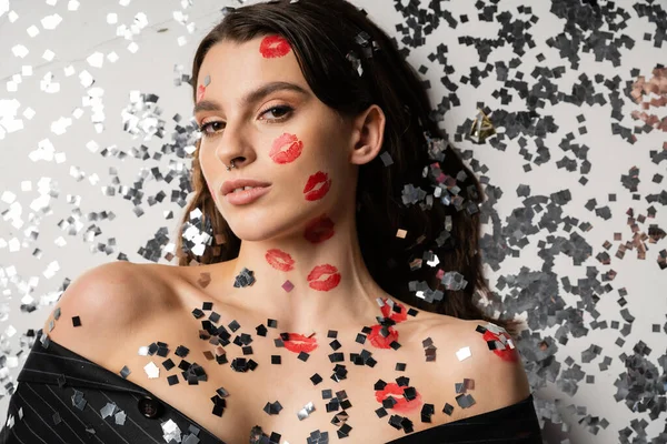 Top view of brunette woman with red kiss prints looking at camera while lying near shiny confetti on grey background — Fotografia de Stock