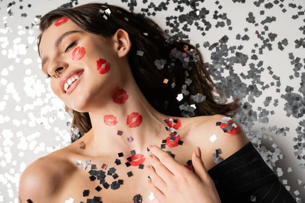 Верхний вид улыбающейся женщины с красными отпечатками губ, лежащих рядом с игристыми серебряными конфетти на сером фоне — стоковое фото